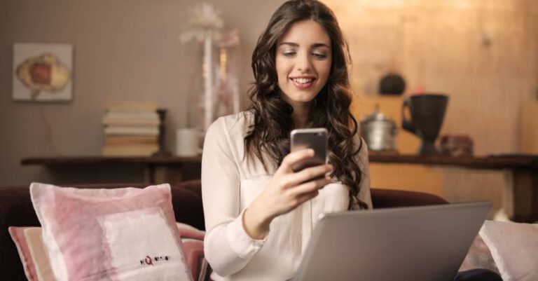 Online - Woman Sitting on Sofa While Looking at Phone With Laptop on Lap